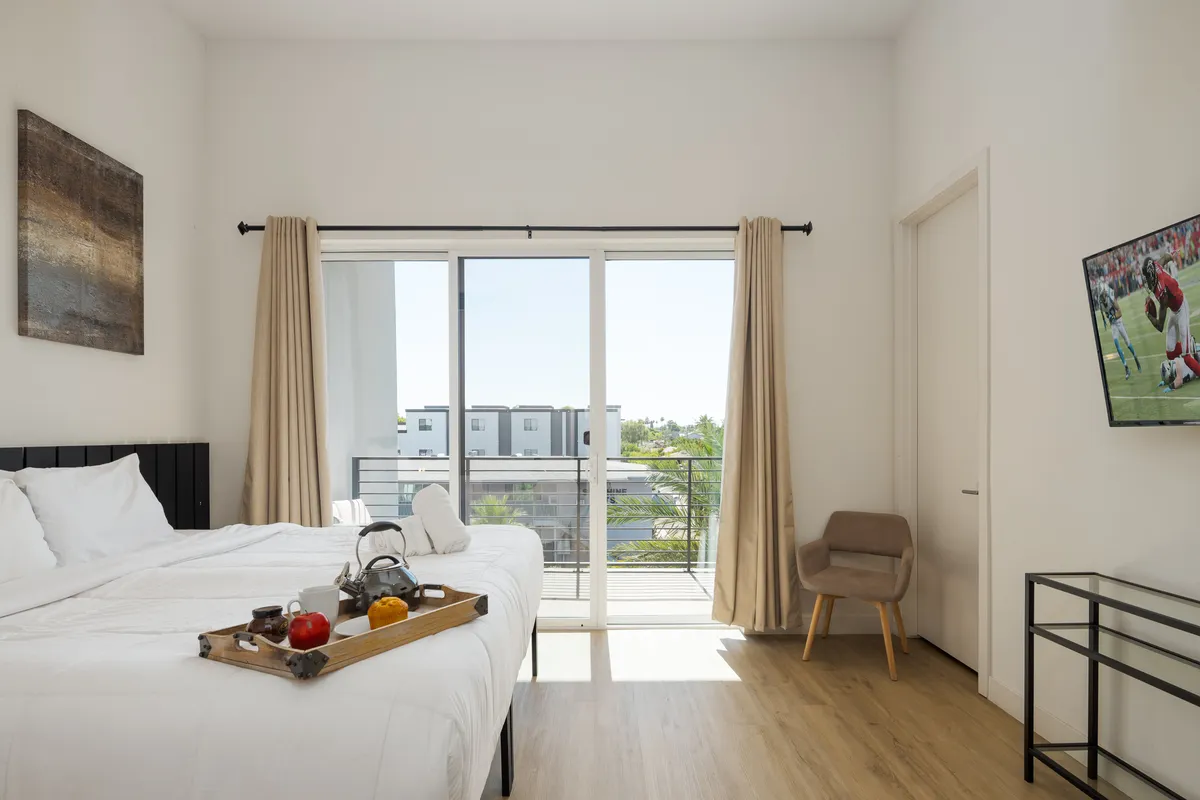 A sunlit bedroom with doors opening to a private balcony, a TV opposite the bed, and a tray with fruit and tea. Natural light fills the space, creating an inviting, comfortable environment ideal for relaxation and enjoying both indoor comforts and outdoor sanctuary.