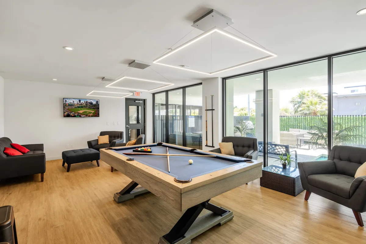 A lively clubhouse interior with a pool table, several comfortable sofas, a television, and large windows letting in natural light. The room is set up for friendly games, conversation, and casual relaxation, embodying the social spirit of the community.