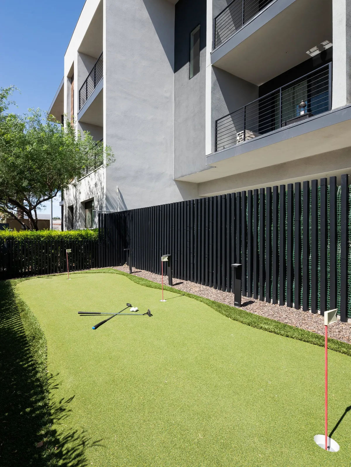 A playful exterior scene featuring a putting green, modern architecture, neatly manicured lawns, and a black fence. The space is designed for both social gatherings and individual practice, set against a sunny backdrop.
