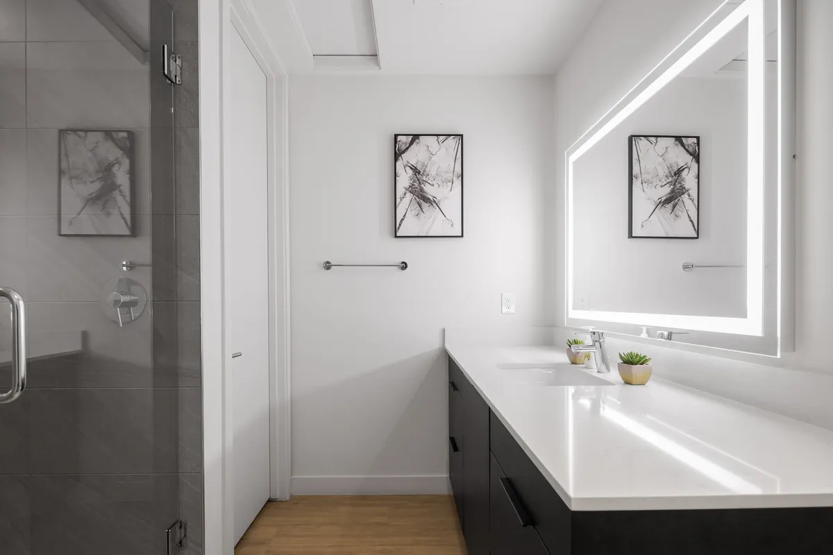 A spa-like ensuite bathroom with a spacious vanity featuring dual sinks, an illuminated mirror, and stylish framed artwork. The setting is modern, tranquil, and designed for peaceful self-care routines.