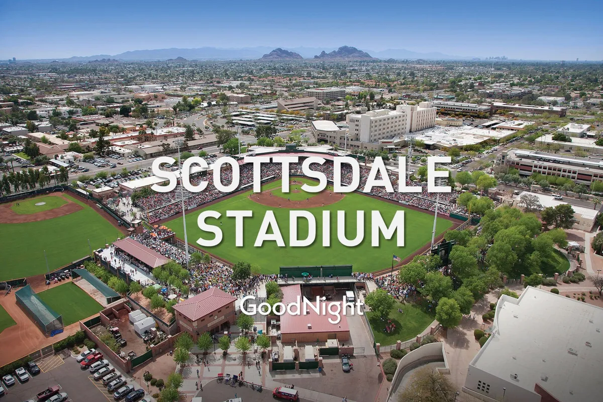 A lively baseball field filled with a crowd of spectators, surrounded by city buildings and a scenic mountain backdrop. The atmosphere is buzzing with excitement, embodying the communal spirit of attending outdoor events and games with friends.