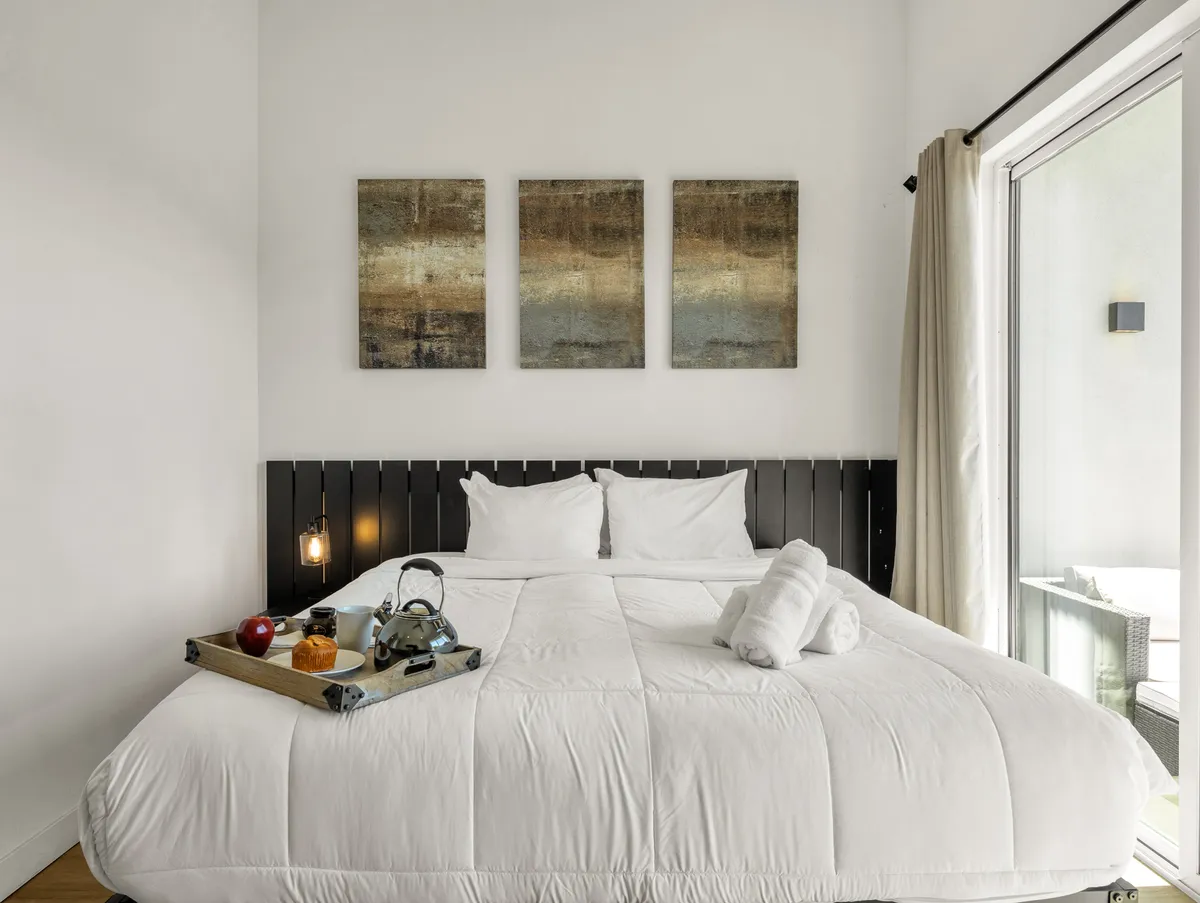 A cozy bedroom featuring a bed with white bedding, calming artwork on the wall, and a tray with breakfast items set atop the bed. The atmosphere exudes comfort and a serene start to the day, with inviting textures and a soft, warm light.