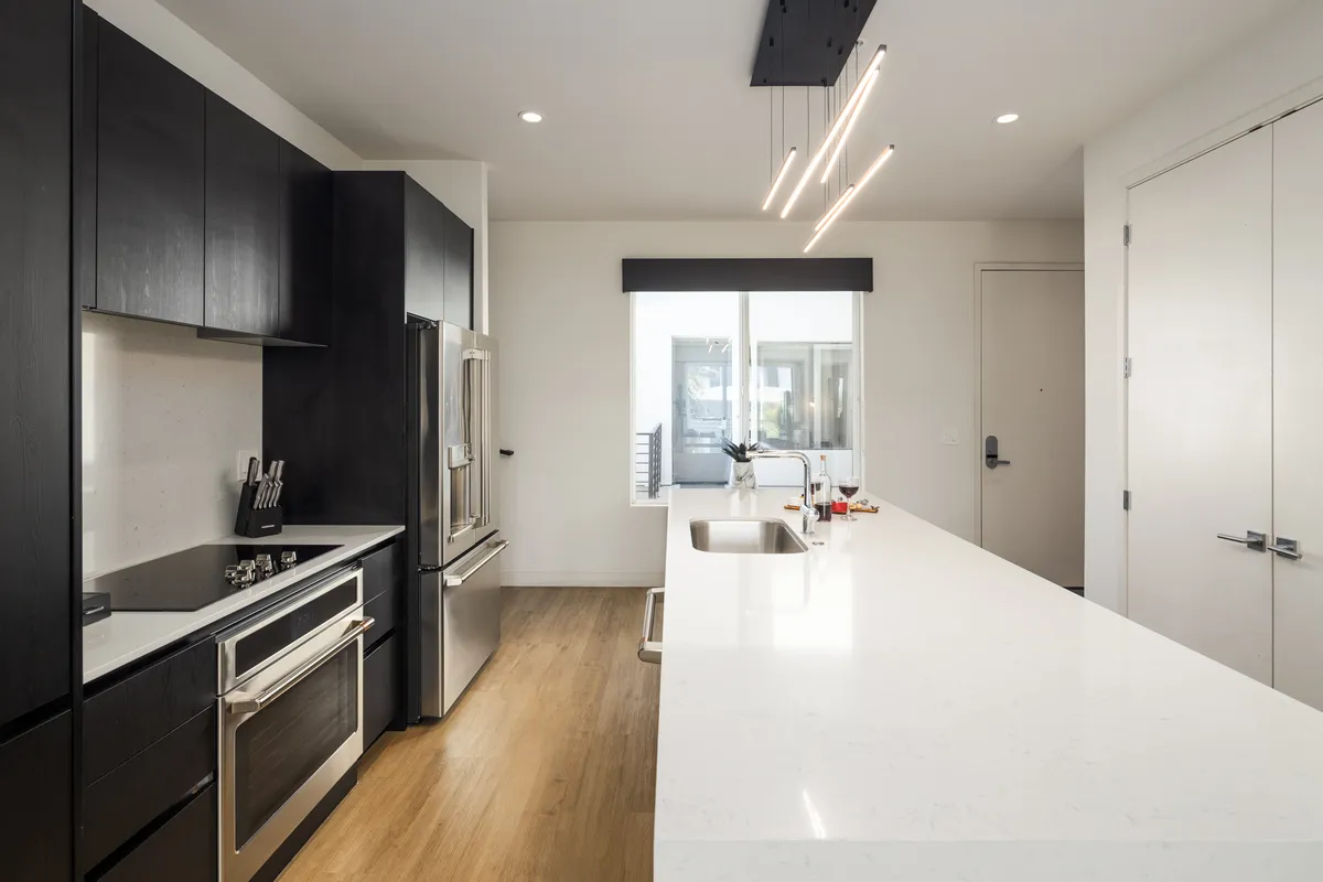 A bright, contemporary kitchen with a sleek central island, stainless steel appliances, and ample natural light flooding in through a large window. The space is open and inviting, highlighting generous counter space and a functional layout ideal for culinary creativity.